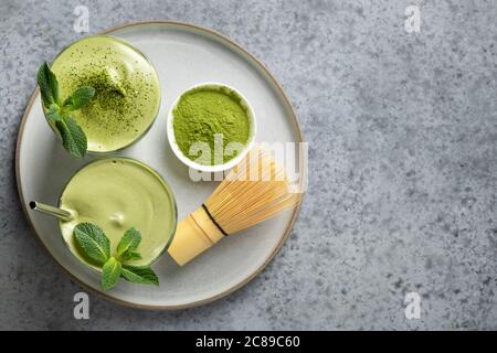 Zwei Gläser Dalgona Matcha Latte Tee, Bambusbesen und Matcha Pulver auf grau. Stockfoto