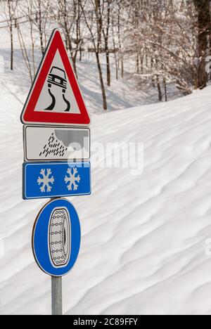 Warnschilder für rutschige Straßen durch Regen oder Eis und obligatorische Schneeketten mit schneebedeckten Hügel im Hintergrund Stockfoto