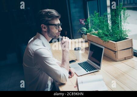 Verträumter Ponder junge Mann in formeller Kleidung sitzt an seinem Arbeitsplatz in einem modernen Coworking im Loft-Stil und schaut denkend ins Fenster Stockfoto