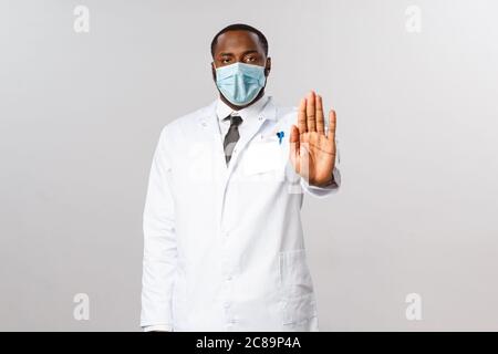 Coronavirus, Kovid19 und Gesundheitskonzept. Der seriöse afroamerikanische Arzt zeigt ein Stoppschild, warnt und unterlässt Patienten, selbst nach draußen zu kommen Stockfoto