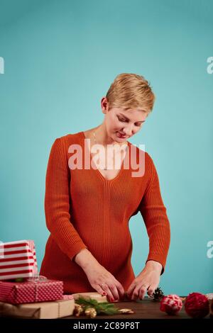 Lächelnde schwangere salerin bereitet Geschenke für Kunden in ihrem Geschenkeladen zur weihnachtszeit vor und macht eine Verbeugung auf der roten Geschenkbox, isoliert im Studio über blauer BA Stockfoto