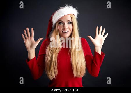 Innenaufnahme der verblüfften blonden langhaarigen Frau in Santa Hut und roten Kleidern, mit den Händen nach oben, Posen gegen schwarze Wand mit leeren Kopie Raum. Stockfoto