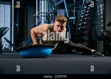 Vorderansicht des muskulösen kaukasischen Mannes Training auf dem Boden mit Balance Halbball. Nahaufnahme des jungen Sportlers Muskeln bauen, tun Liegestützen auf einer Hand in leeren Fitnessraum. Konzept des Bodybuilding. Stockfoto