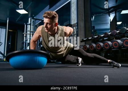 Vorderansicht der muskulösen kaukasischen Mann tun Liegestützen auf einer Hand auf dem Boden mit Balance Halbball. Nahaufnahme des jungen Sportlers, der in einer leeren Turnhalle Muskeln baut. Konzept von Bodybuilding, Sport. Stockfoto