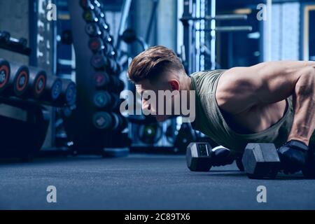 Crop von muskulösen kaukasischen Mann Training auf dem Boden mit Humbbels im Sportverein. Nahaufnahme des jungen Sportlers Muskeln bauen, tun Liegestütze in leeren Turnhalle, blaues Licht. Konzept des Sports, Bodybuilding. Stockfoto