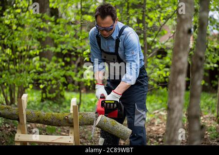 Brutal erwachsenen Mann in Overalls Sägen Baumstamm mit Kettensäge auf Sawhorses. Stockfoto