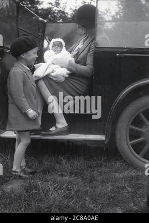 1930er Jahre, historisch, eine Mutter hält ihr Kleinkind, während sie auf der Beifahrerseite eines offenen Autos der Ära sitzt, mit ihrem jungen Sohn aussehen Phantasie in seiner Jacke und Baskenmütze, neben dem Fahrzeug stehend, lächelnd, England, Großbritannien. Stockfoto