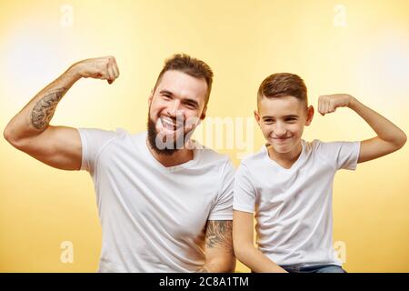 Fröhlicher bärtiger Vater und Sohn zeigen Bizeps und lächeln an der Kamera isoliert auf gelb Stockfoto