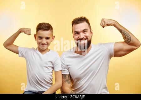 Fröhlicher bärtiger Vater und Sohn zeigen Bizeps und lächeln an der Kamera isoliert auf gelb Stockfoto