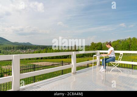 Konzentrierte stilvolle Frau sitzt auf dem weißen Balkon der Terrasse in einem Laptop-Apartment und arbeitet, starrte aufmerksam auf den Bildschirm. Schönes Mädchen Stockfoto