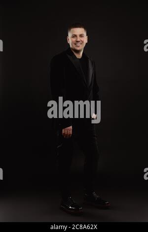 In voller Länge Porträt eines jungen männlichen Erwachsenen in einem eleganten Anzug posiert mit seiner Hand in der Tasche isoliert auf schwarzem Hintergrund Stockfoto