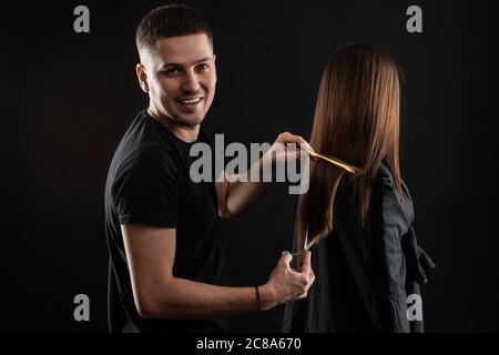 Schöner junger Friseur lächelt an der Kamera, während Haare kämmen, um schöne junge Frau im Schönheitssalon Stockfoto