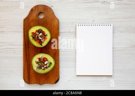 Hausgemachter Pico de Gallo gestopft Avocado auf einem rustikalen Holzbrett, leerer Notizblock auf einer weißen Holzfläche, Draufsicht. Flach, über Kopf, von oben. Sp Stockfoto