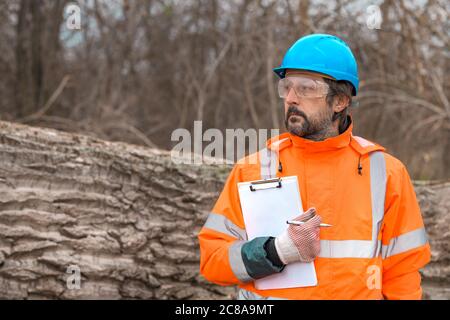 Forsttechniker, der mit einem Notizblock in der Zwischenablage neben einem Baumlogan in der Walde posiert, ist zuversichtlich, dass männliche Fachmann Daten auf dem Feld sammelt Stockfoto