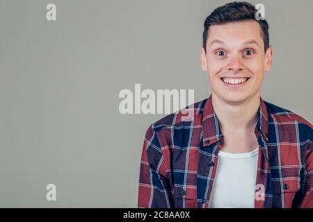 Hübscher junger Mann auf grauem Hintergrund Blick in die Kamera. Porträt des Lachens jungen Mannes mit Händen in den Taschen, graue Wand gelehnt. Glücklicher Kerl smi Stockfoto