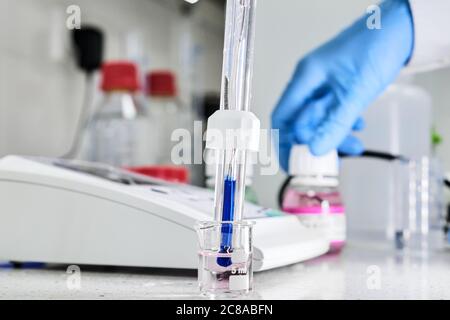 Frau in einem Gummihandschuh mit einer Glaselektrode zur Messung des pH-Wertes der Lösung mit einem pH-Messgerät. Labor für analytische oder elektrochemische Untersuchungen. Stockfoto