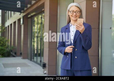 Portrait einer attraktiven reifen Geschäftsfrau, die klassischen Anzug und Brillen mit Mobiltelefon trägt und lächelt, während sie im Freien gegen das Bürogebäude steht. Geschäftsleute Stockfoto