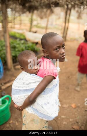 Joan Nkinzi (10) trägt ihren kleinen Bruder Luice Ssekalala (12 Monate) auf dem Rücken in ihrem Zuhause im Masaka District, Uganda. Beide sind Kinder von VEA J Stockfoto