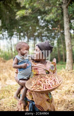 Eine lächelnde kleine Bäuerin hält ihren Enkel und einen Korb mit ihrer frisch geernteten Bohnenernte auf ihrer Farm im ländlichen Lyantonde District in Uganda. Stockfoto