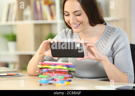 Glückliche Frau, die mit Blitz auf dem Smartphone fotografiert, die zu Hause auf einem Schreibtisch sitzt Stockfoto