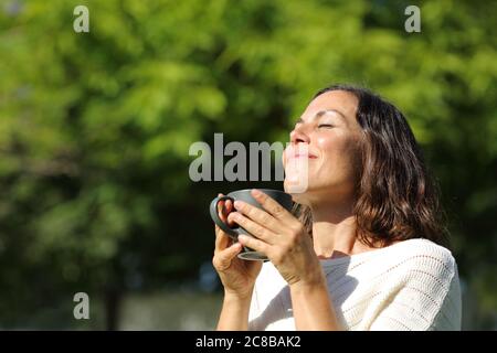 Zufriedene Erwachsene Frau riecht Kaffee Tasse steht in einem grünen Park im Sommer Stockfoto