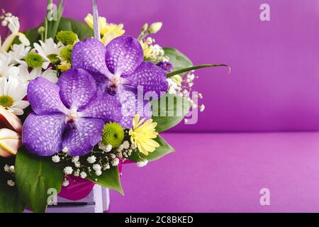 Schöner Blumenstrauß in lila Korb auf lila Hintergrund mit Kopierer Platz. Orchideen, Lilien, Chamomilen florale floristische Komposition. Begrüßung Stockfoto