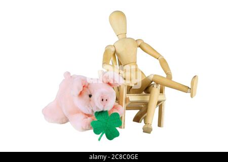 Eine Holzpuppe sitzt auf einem Stuhl neben einem Plüschpiggy mit Kleeblatt über Weiß Stockfoto