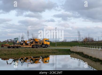 Tieflader gesehen Transport von Baufahrzeugen aus der großen Wohnsiedlung mit einem Landschaftsteich und Steinbrücke im Vordergrund. Stockfoto