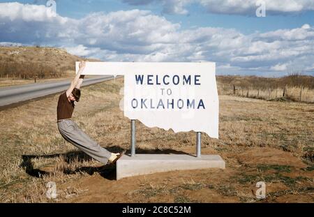 Ein junges Mädchen schwingt aus dem Ende eines Schildes an der Staatsgrenze von Oklahoma, USA in den 1950er Jahren. Das Schild begrüßt die Menschen im Staat und das Metall ist in der Umrandung seiner Grenze ausgeschnitten. Stockfoto