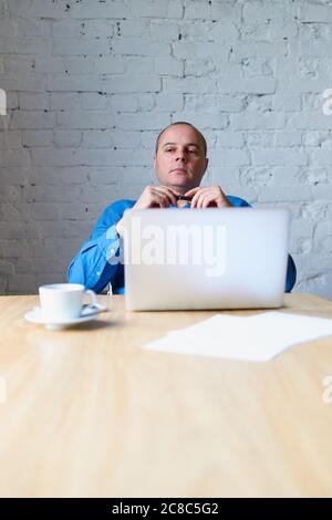 Schöner schöner, reifer Mann, der vor dem Laptop am Tisch sitzt und dem Untergebenen zuhört. Mann mit legerer Kleidung in blauem Hemd im Büro Stockfoto