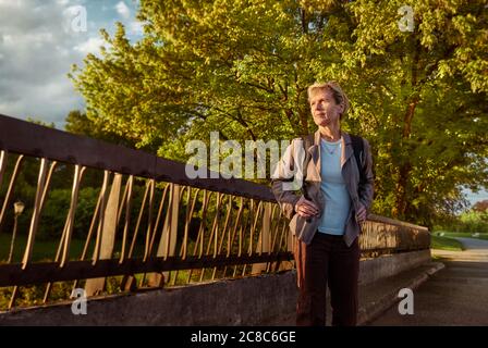 Eine Frau mittleren Alters steht auf einer Brücke vor dem Parkeingang und bewundert den Sonnenuntergang. Stockfoto