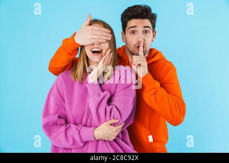Bild eines Mannes, der die Augen seiner Freundin bedeckt, während er eine Stille zeigt, die isoliert auf blauem Wandhintergrund liegt. Stockfoto