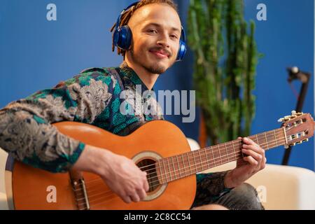 Junger Mann mit Kopfhörern sitzt auf dem Sofa und spielt Gitarre Stockfoto