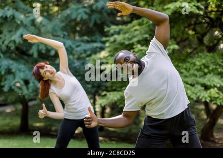 Kaukasische Frau und afrikanischer Mann, Fitness-Paar, Training zusammen Stretching Körperübungen im Park bei Sonnenaufgang, lächeln, Kamera und Stockfoto