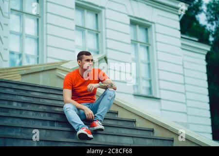 Ein junger Mann in einem roten T-Shirt und Jeans posiert auf der Treppe des Anwesens. Ein erwachsener Mann in roten Turnschuhen sitzt auf den Stufen des Palastes. Stockfoto