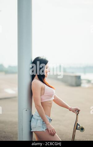 Urban modische Mädchen mit Longboard posiert im Freien auf dem Weg zum Sonnenuntergang. Stockfoto