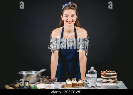 Happy Pretty Konditorei Köchin Frau in Cutaway Kleid und Schürze hinter Tisch mit frisch gebackenen Desserts und Küchengeräte auf blauem Hintergrund stehen Stockfoto