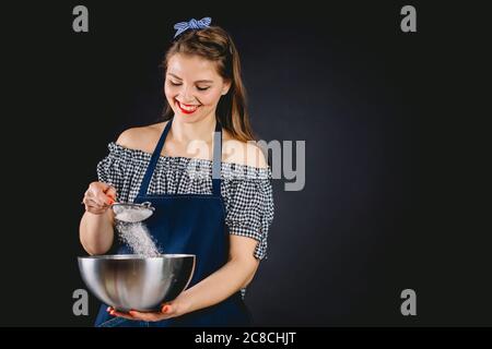 Glückliche Hausfrau in dunkelblauer Schürze durchsiebt ein Sieb auf schwarzem Hintergrund im Studio Stockfoto