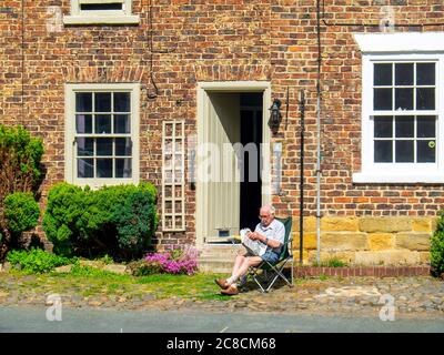 Ein älterer Herr, der vor seinem Reihenhaus am Fluss Leven im Stadtzentrum von Stokesley, North Yorkshire, seine Zeitung liest Stockfoto