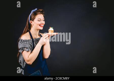 Schöne Konditorin Frau genießen frisch gebackenen Deliciuos Cupcake in den Händen, zeigt Bewunderung auf ihrem Gesicht Stockfoto