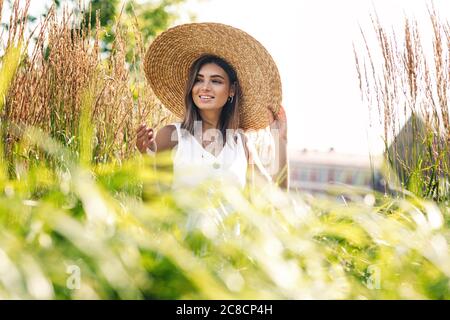 Junge glückliche Frau in großen Strohhut auf dem Feld stehend und wegschauen. Stilvolle weibliche Wandern im Freien. Stockfoto