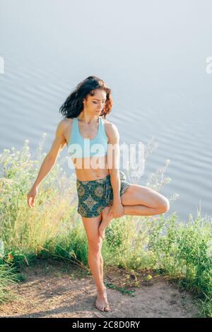 Schöne lächelnde glückliche junge Frau, die draußen arbeitet, barfuß Yoga macht, in Vrksasana-Haltung steht, Baumhaltung, volle Länge Stockfoto