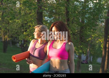 Zwei Frauen, die während des Sonnenuntergangs mit einer Yogamatte auf einem ländlichen Gebiet in Sportkleidung und Yoga unterwegs sind Stockfoto