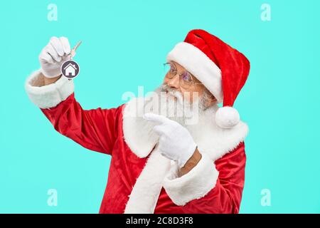 Vorderansicht eines lächelnden alten Mannes in rotem Kostüm mit Schlüsseln und Hausschild. Isoliertes Porträt des Weihnachtsmanns, der auf Schlüssel in der Hand auf blauem Studiohintergrund zeigt. Konzept der Ferien. Stockfoto