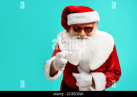 Seitenansicht des lächelnden Mannes im roten Weihnachtsmann Kostüm. Isoliertes Porträt eines älteren Mannes mit weißem Bart in einer Sonnenbrille, die auf die Kamera auf blauem Studiohintergrund zeigt. Konzept der Ferien. Stockfoto
