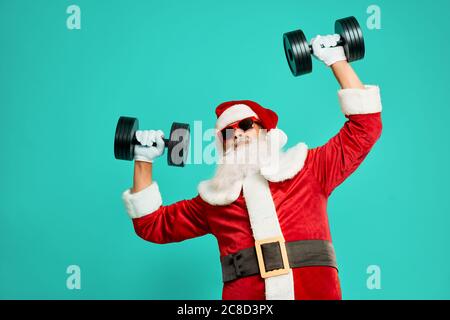 Vorderansicht des sportlichen Weihnachtsmannes mit Hanteln. Isolierte Ernte von lustigen älteren Mann in weihnachtskostüm und Sonnenbrille posiert auf blauem Studio Hintergrund. Konzept von Urlaub, Spaß. Stockfoto