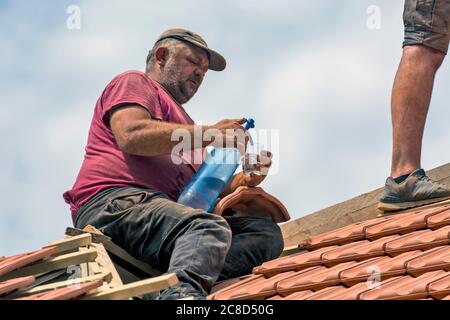 Zrenjanin, Serbien, 23. Juli 2020. Meister arbeiten auf dem Dach eines Privathauses, um eine alte Fliese zu ersetzen. Einer der Meister erfrischt sich mit Stockfoto