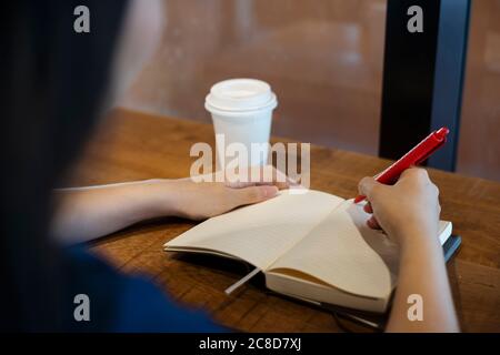 Eine Frau, die im Café mit heißem Kaffee sitzt, das Notizbuch öffnet und ihr Tagebuch schreibt, während sie auf ihren Geliebten als Termin wartet, über die Schulter geschossen Stockfoto