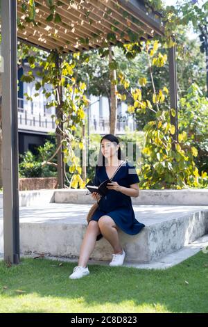Lange Haare Frau auf der Steinbühne sitzen, das Buch lesen, während die Musik mit Kopfhörer hören, ihr Freund nicht rechtzeitig zu erscheinen. Stockfoto