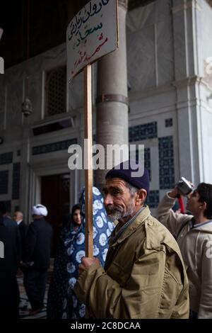 Damaskus, Syrien 03/28/2010: Ein alter bärtiger Mann mit Regenmantel und Wintermütze hält eine Richtplakat auf einem Stock, um vi anzuziehen Stockfoto